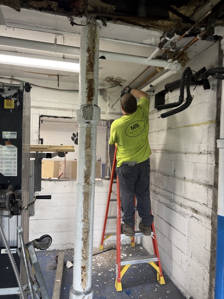 A man in a yellow shirt and pants climbs a ladder against a brick wall, working on a ceiling pipe.