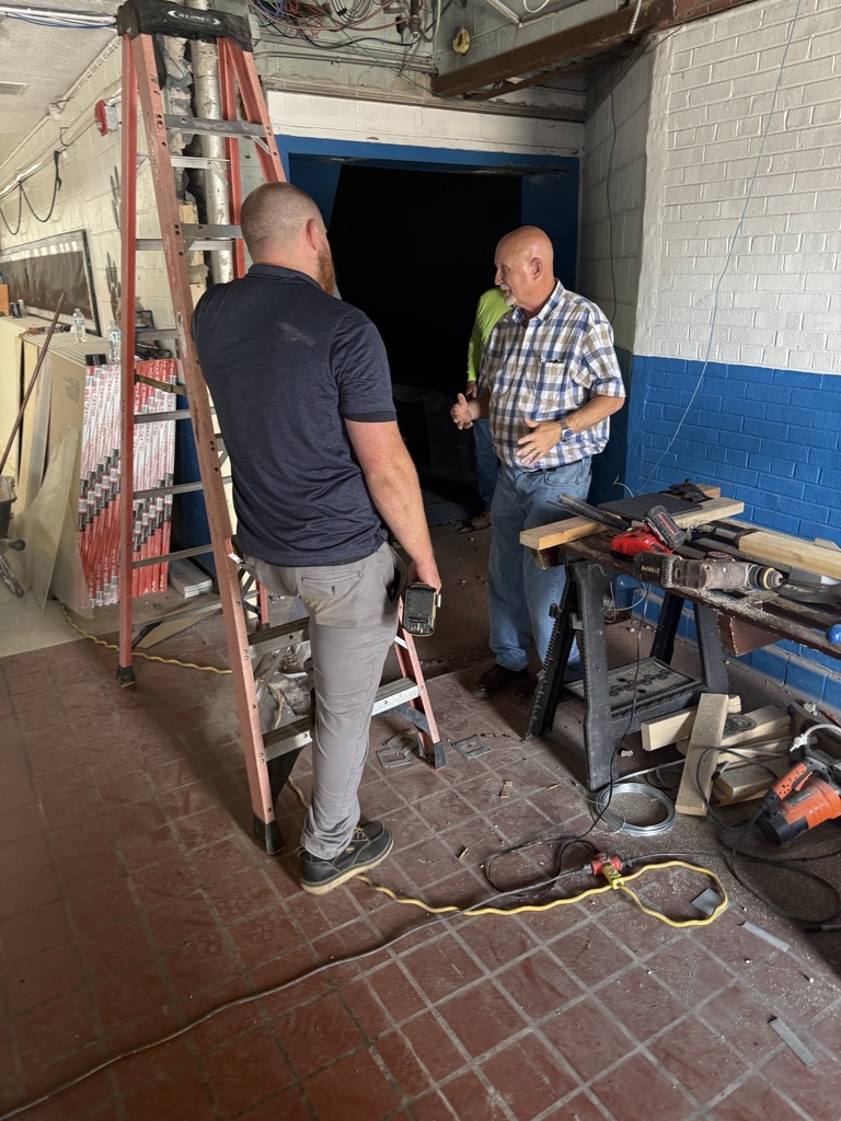 Two men in a room with red brick floors. One stands on a ladder, the other stands by a table.