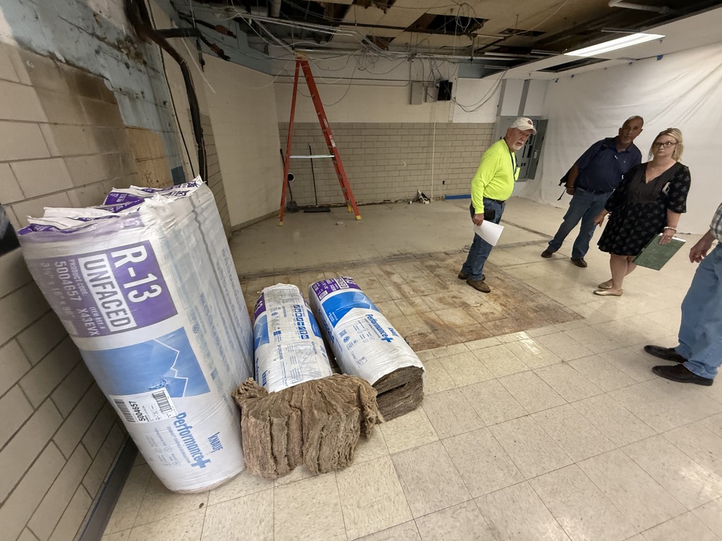 Four people stand in a room under construction with exposed ceiling and walls, next to rolls of flooring material.