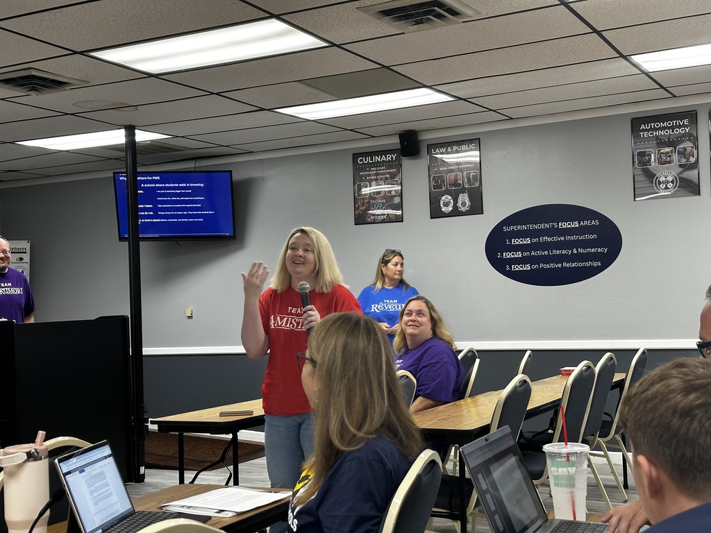 A woman speaks in front of others in a room with tables, laptops, and a monitor on the wall.