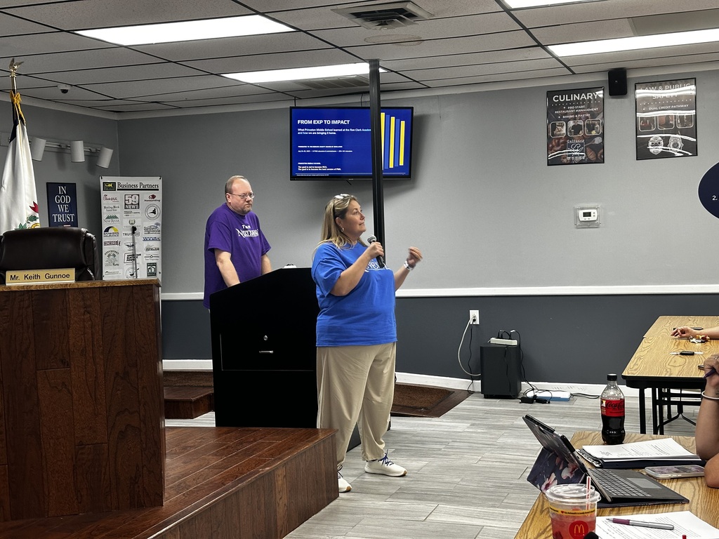 A woman stands at a podium speaking to a group in a room with a monitor and flag.