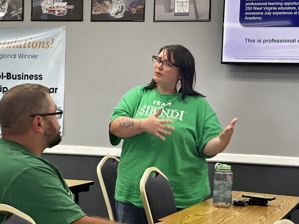 A woman in a green T-shirt explains to a man in a chair at a desk in a classroom.