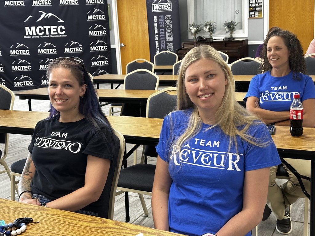 Three women in matching t-shirts sit at a table in a room with chairs and desks.