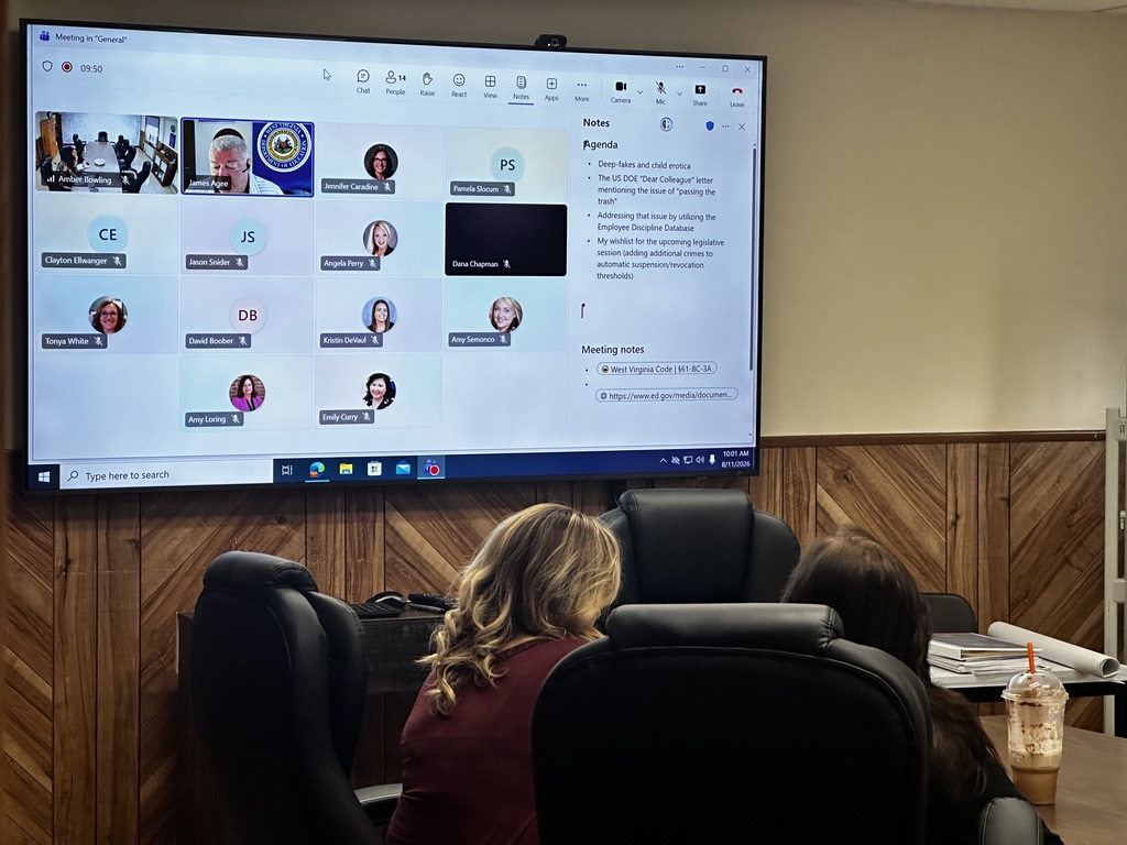 People sit in chairs in a meeting room, watching a presentation on a large screen.