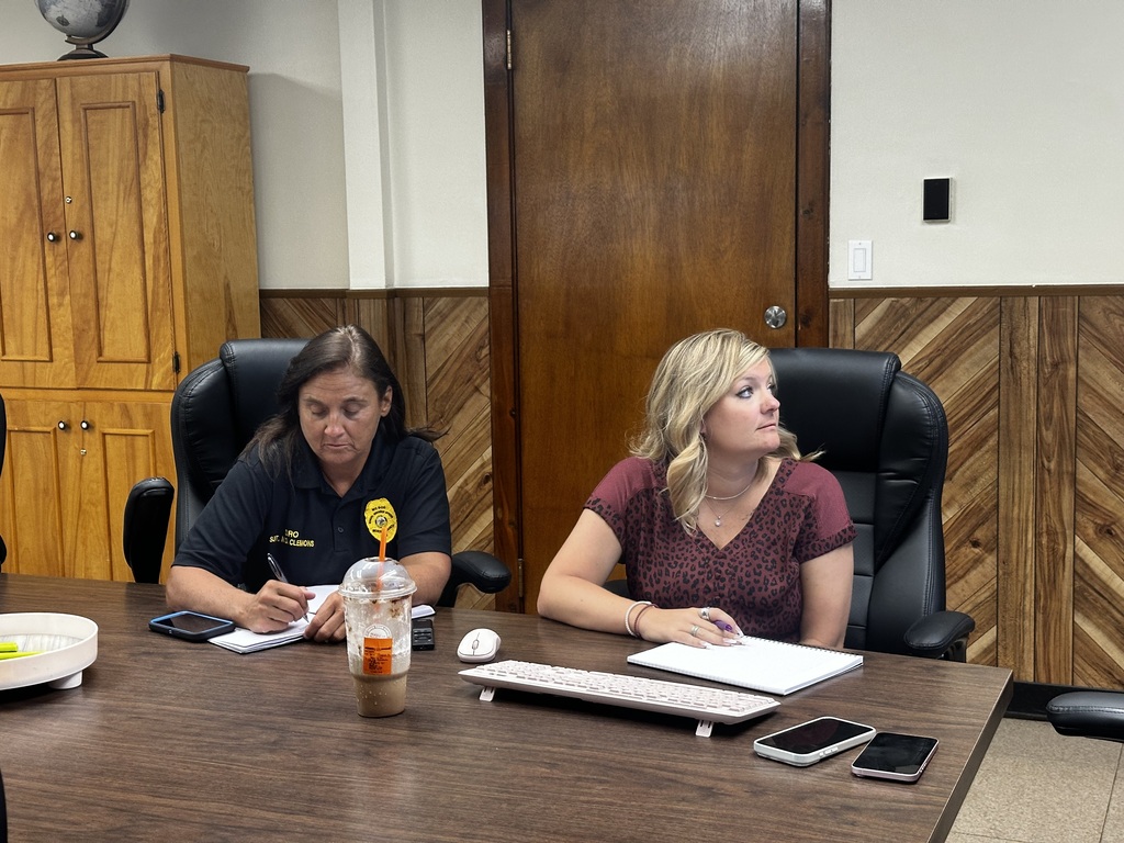 Two women sit at a table, each holding a pen and paper. A laptop, keyboard, and cellphone are on the table.