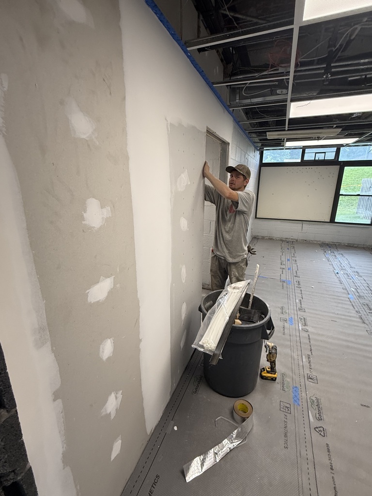 A man is painting a wall in a room, with patches of unfinished areas and a bucket nearby.