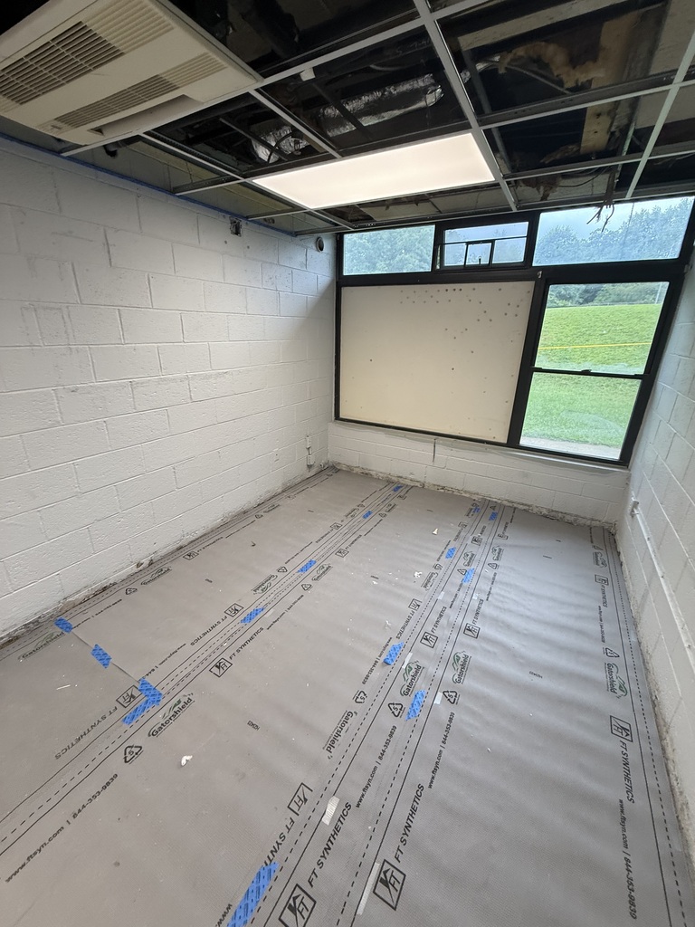 An empty room with white walls and ceiling. The floor is covered in a protective sheet. A window lets in natural light.