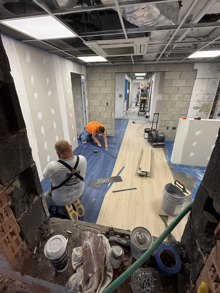 A room under renovation with a new floor being installed. Two workers are present, one on the ground and one on a ladder.