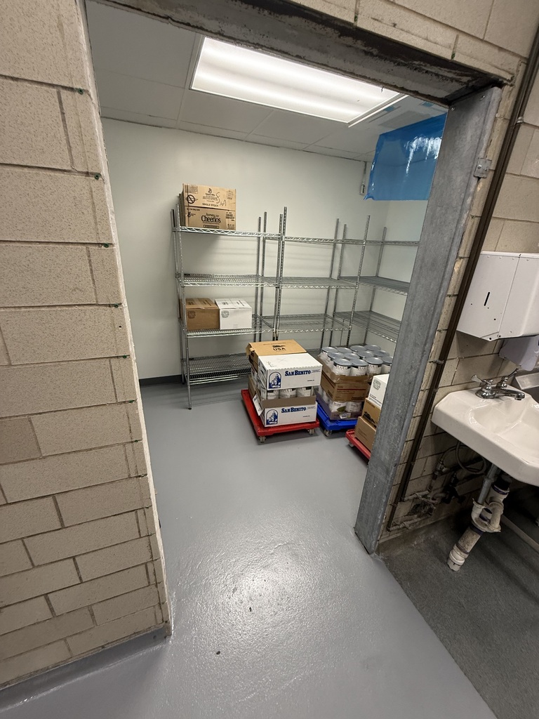 Interior of a small room with white floor, brick wall, metal shelving with boxes, a white sink, and a blue tarp.