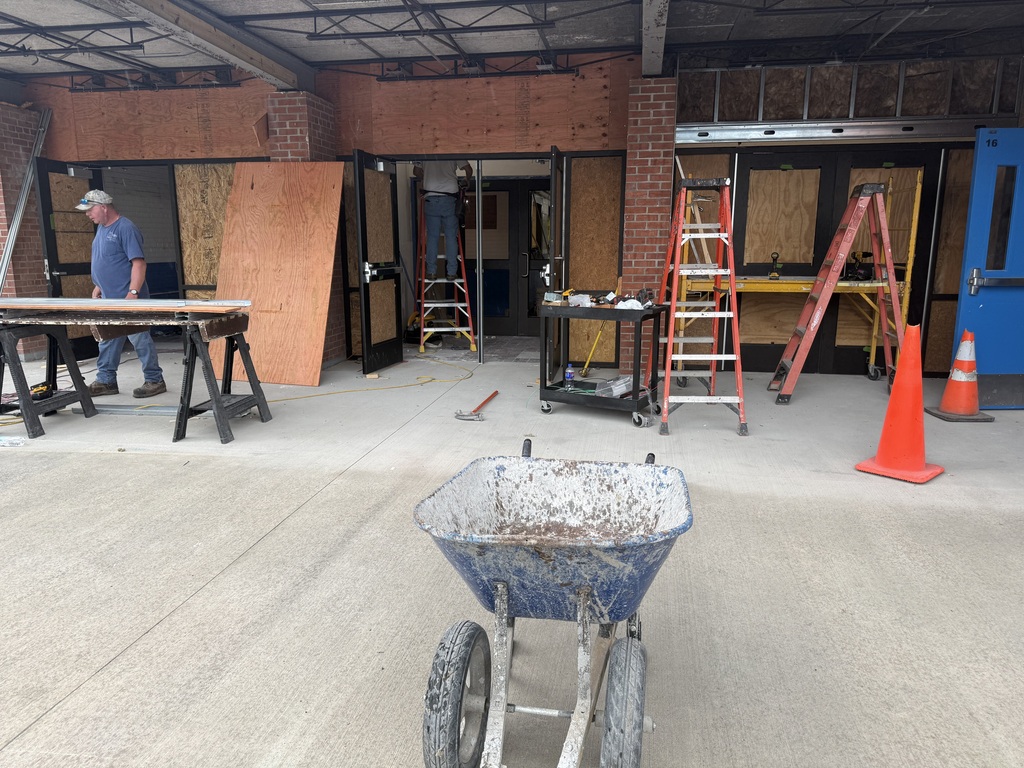 A construction site with a man, ladders, a wheelbarrow, and orange cones on a cement floor.