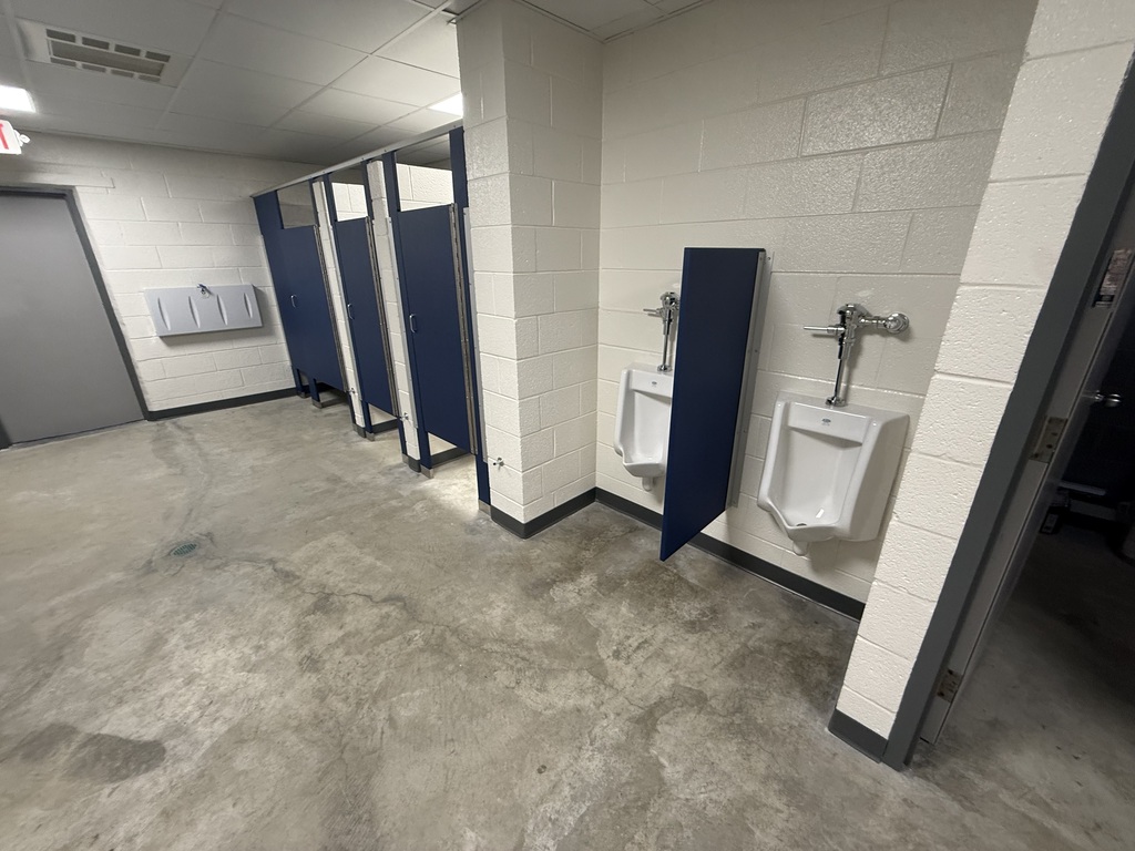 Empty restroom with white walls, gray floor, three blue dividers, two urinals, and a closed gray door.