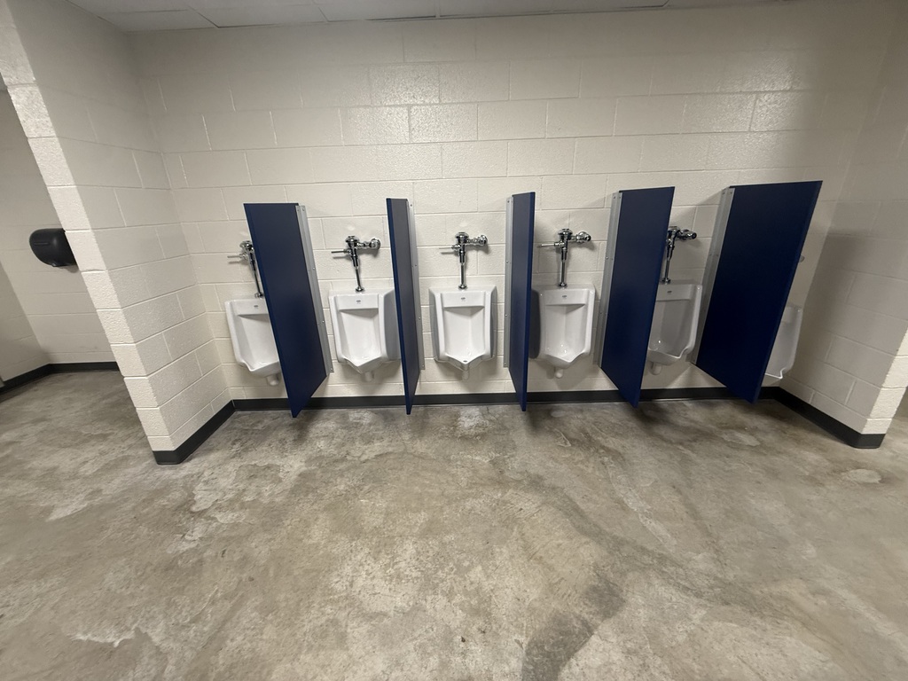 Four blue urinals line a wall in an empty bathroom. The floor is concrete with a gray finish.