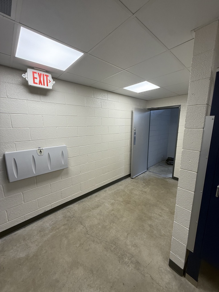 A corridor with white walls and a beige floor. An open door leads to another space. An exit sign is mounted on the wall.