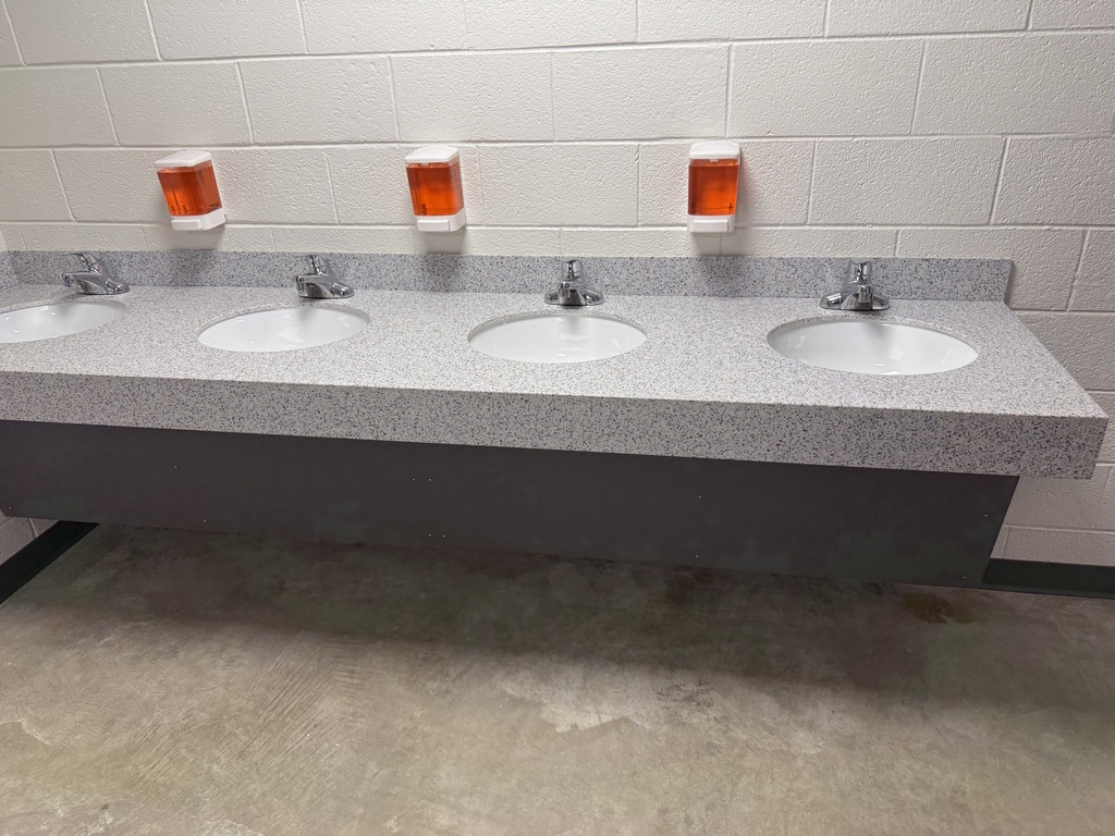 Three sinks on a gray countertop in a bathroom. Above each sink is an orange soap dispenser.