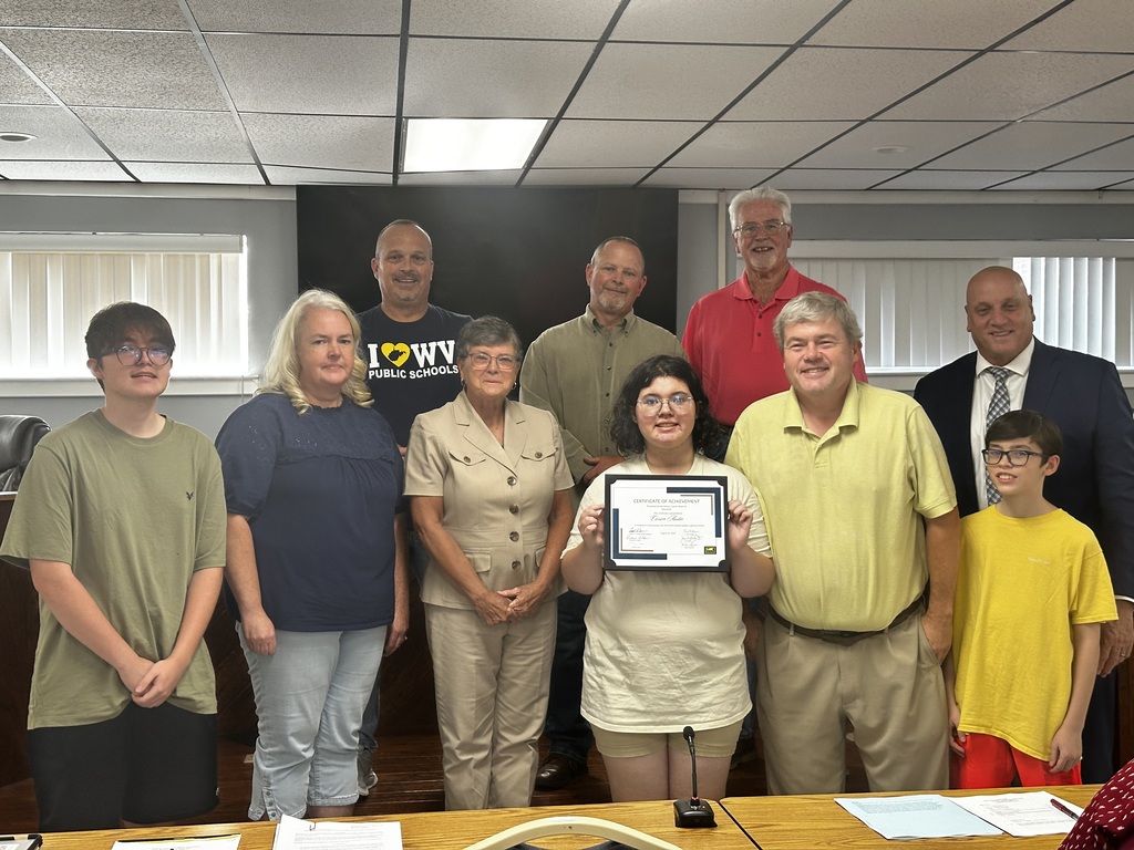 Group of adults and children stand in a room; woman in front holds a certificate.