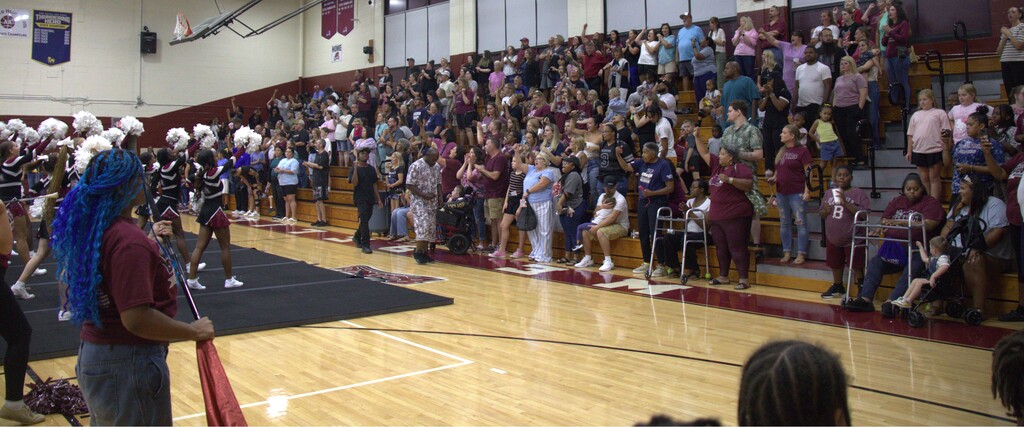 Many people watching a performance on a basketball court, some in maroon, others in white, with an audience on bleachers.