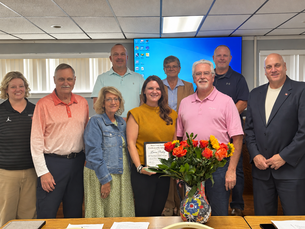 A group of people stands around a table with a flower arrangement and certificate. A woman in the center holds the certificate.