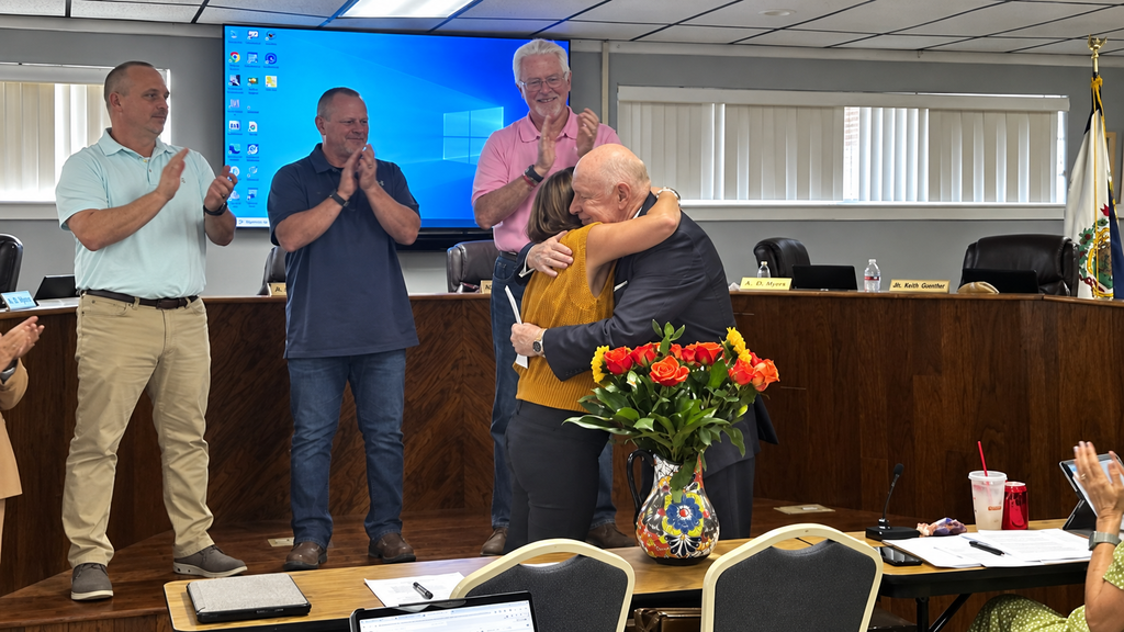 Group of people in a room. A man hugs a woman. A podium is in the foreground.