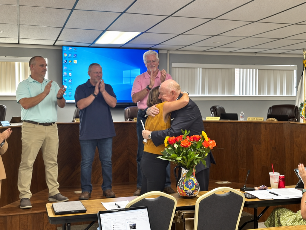 A group of people in a meeting room. A man and woman hug while others clap. Desks with laptops, chairs, and a table with flowers are visible.