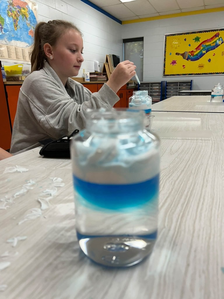 A girl in a classroom sits at a desk, adding liquid to a jar with a white and blue layered effect.