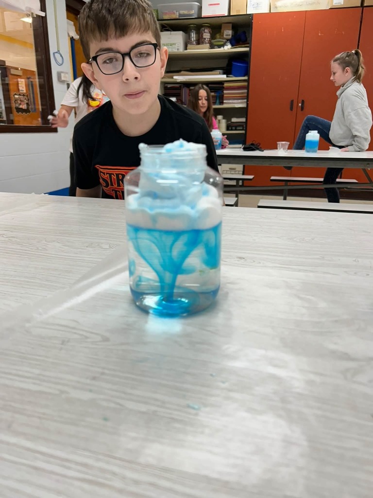 A child with glasses examines a bottle of blue liquid on a table in a classroom.