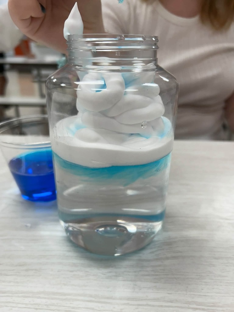 A person adds whipped cream to a jar of water on a table. Layers of foam and water.