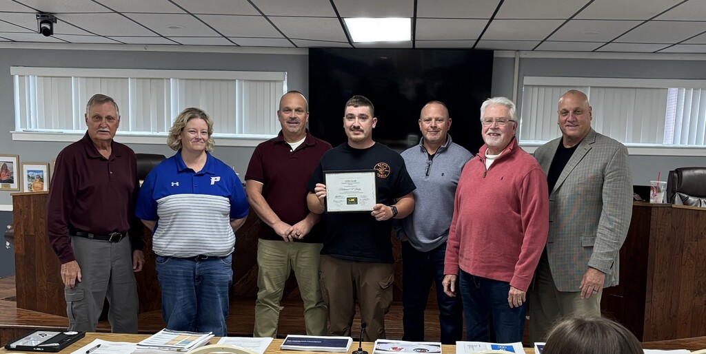 A group of six adults stand around a table in an office, with one person holding a framed certificate.