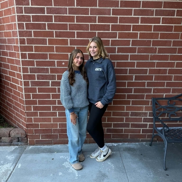 Two individuals, one in a blue sweater and the other in a gray sweater, stand next to a brick wall.
