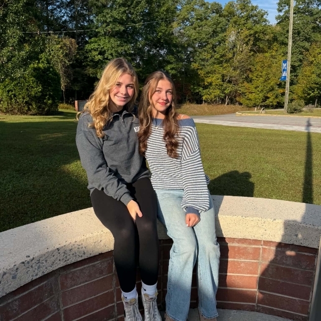 Two people sitting on a curved brick wall, smiling and posing for a photo. Behind them is a grassy area with trees.