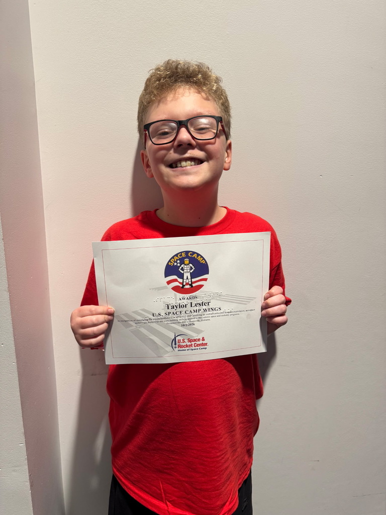 Boy in a red shirt and glasses holding a certificate with a logo. White wall behind him.