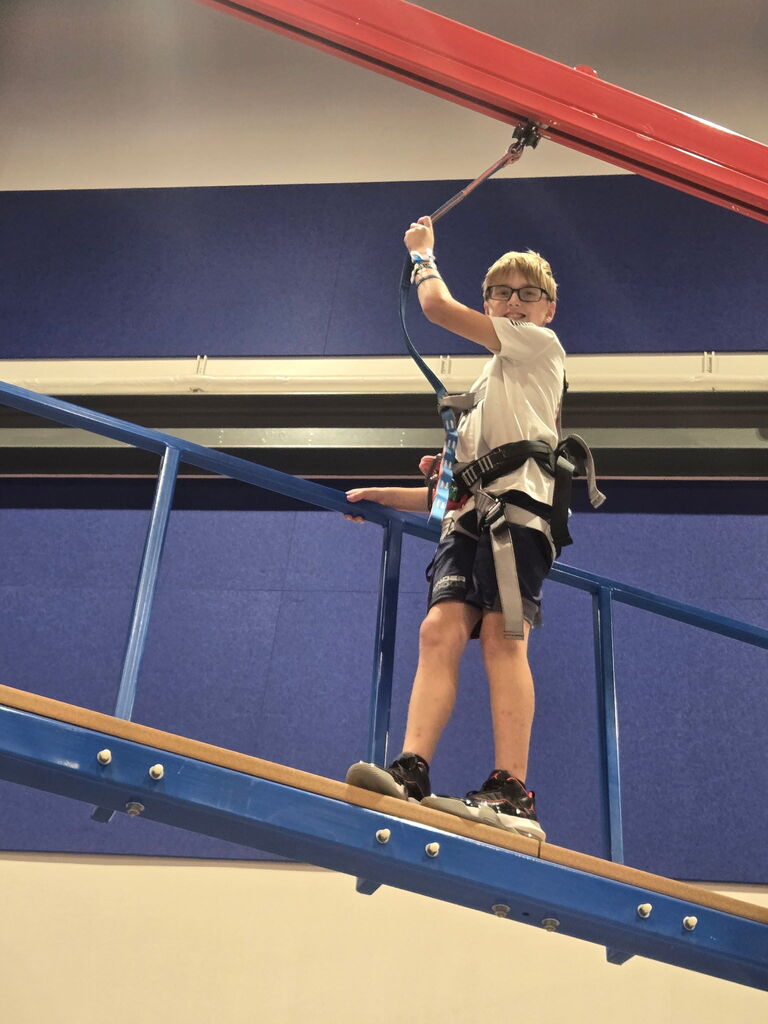 A child wearing a white T-shirt, glasses, and sneakers stands on a wooden platform attached to a red metal beam.