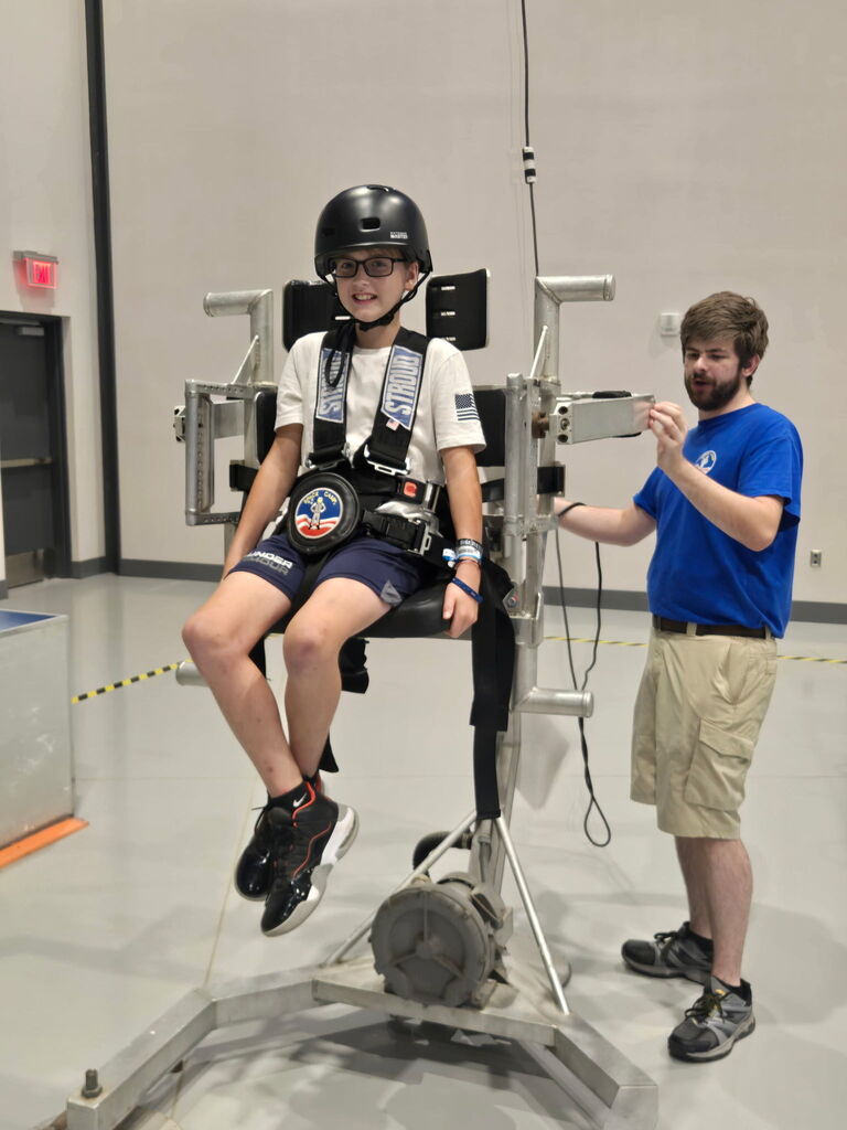 Person in harness on chair. Another person holds equipment. Indoor room with white walls and floor.