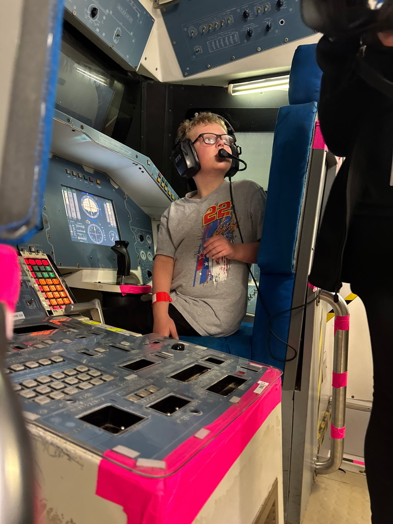 A child wearing glasses and headphones sits at an electronic control panel inside a cockpit.