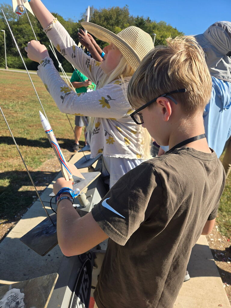 Two children are examining a small rocket on a platform. One wears a straw hat and glasses.