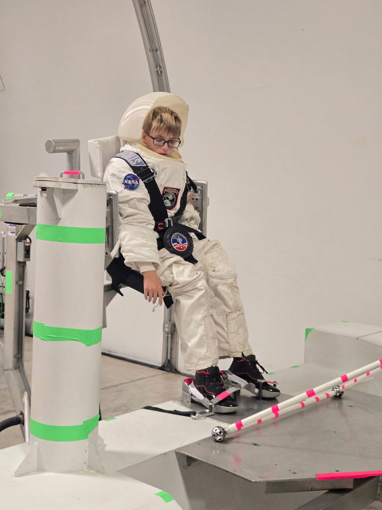 A person wearing a space suit sits on a white chair in a room. The suit has NASA insignia.