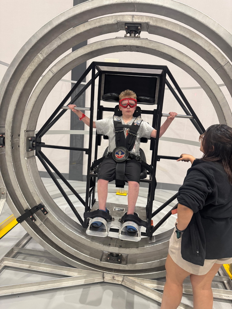 A person in a wheelchair inside a large, circular, silver ride. A woman in black holds the ride's handles.