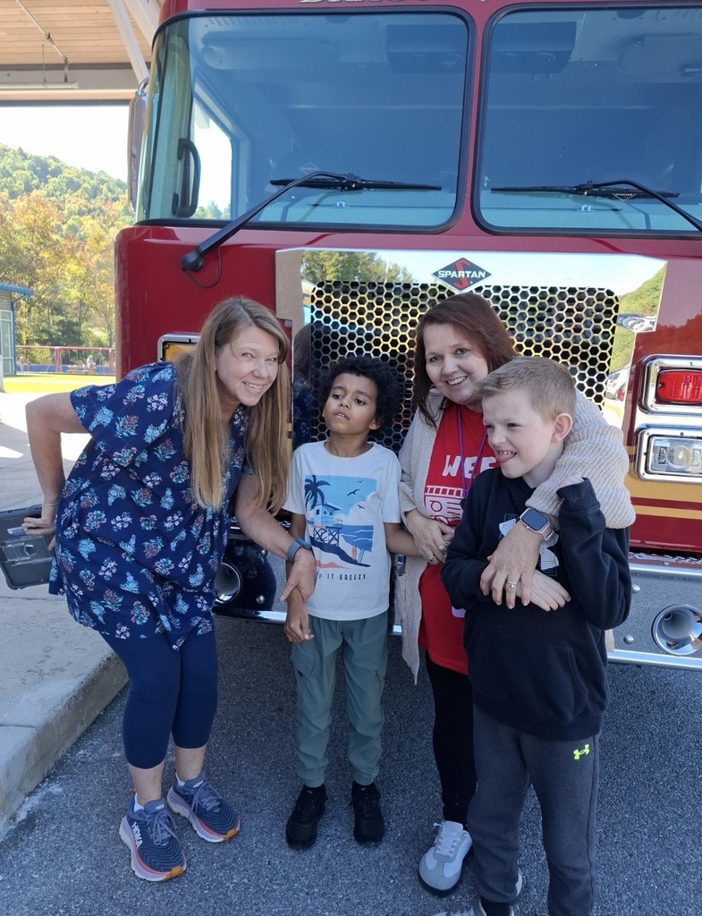 Five individuals stand in front of a fire truck, smiling. A woman in blue and two children wear sneakers.