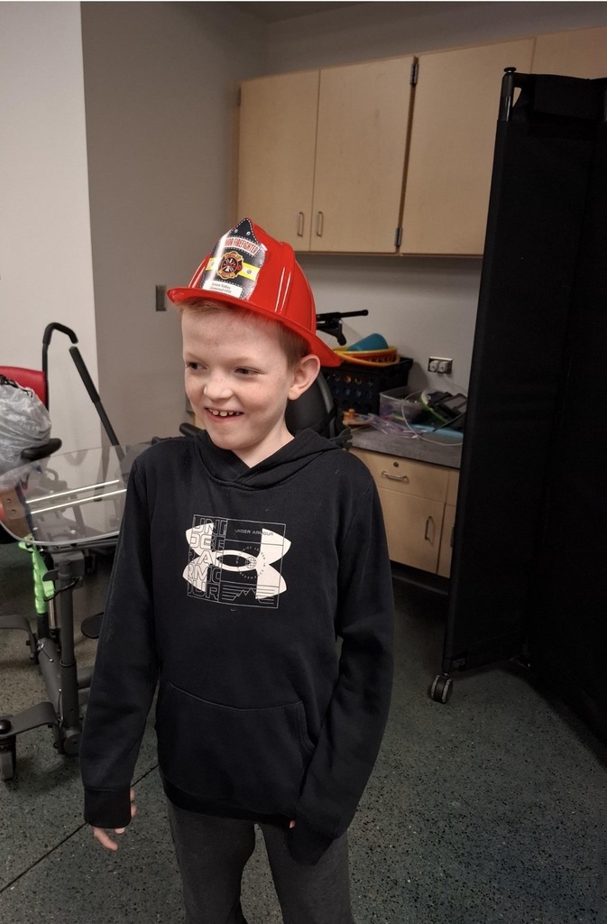 A boy with a red firefighter hat smiles, standing in a room with cabinets, a trolley, and a black screen.