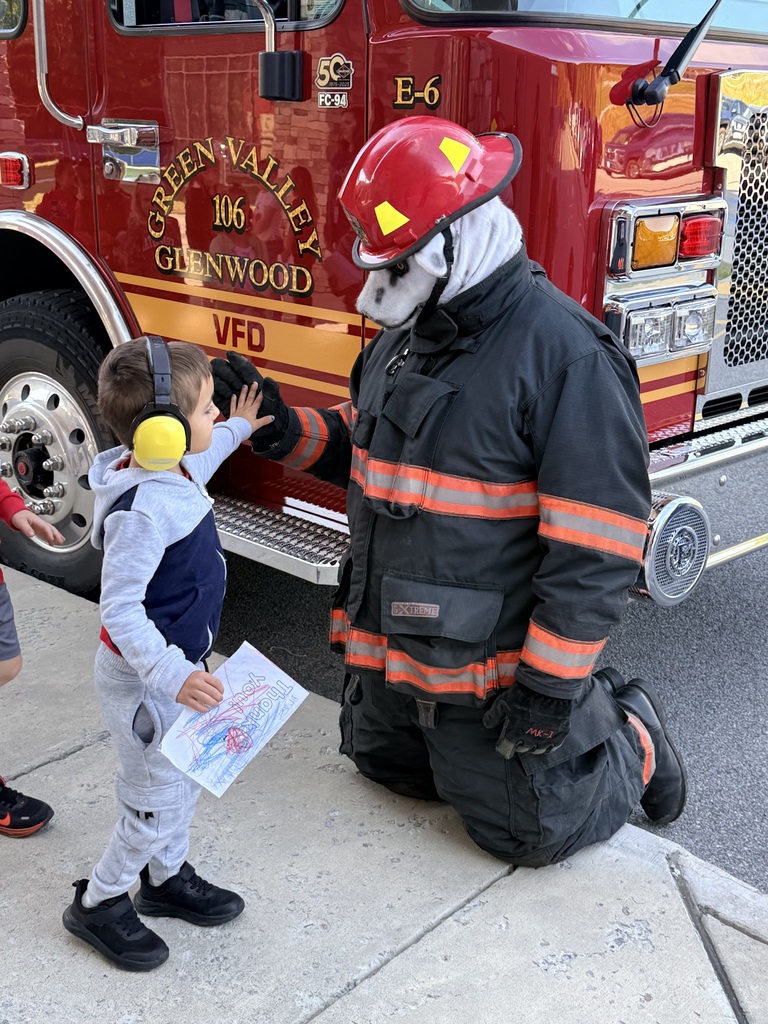 A firefighter crouches to speak with a child. The child holds paper, and a red fire truck is behind them.