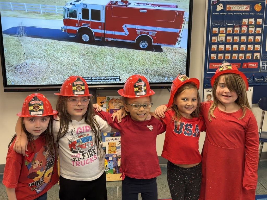 Five children wearing red fireman's hats stand next to each other in front of a large screen.