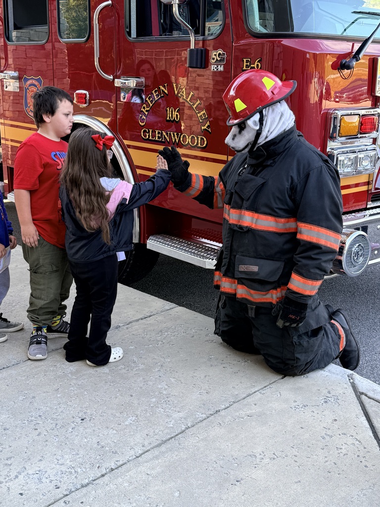 A firefighter in uniform kneels, shaking a young girl's hand. Behind them, a fire engine is parked.