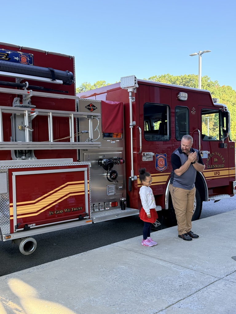 A man and a child stand in front of a red fire truck, the man speaking. Behind them, a streetlight and trees are visible.