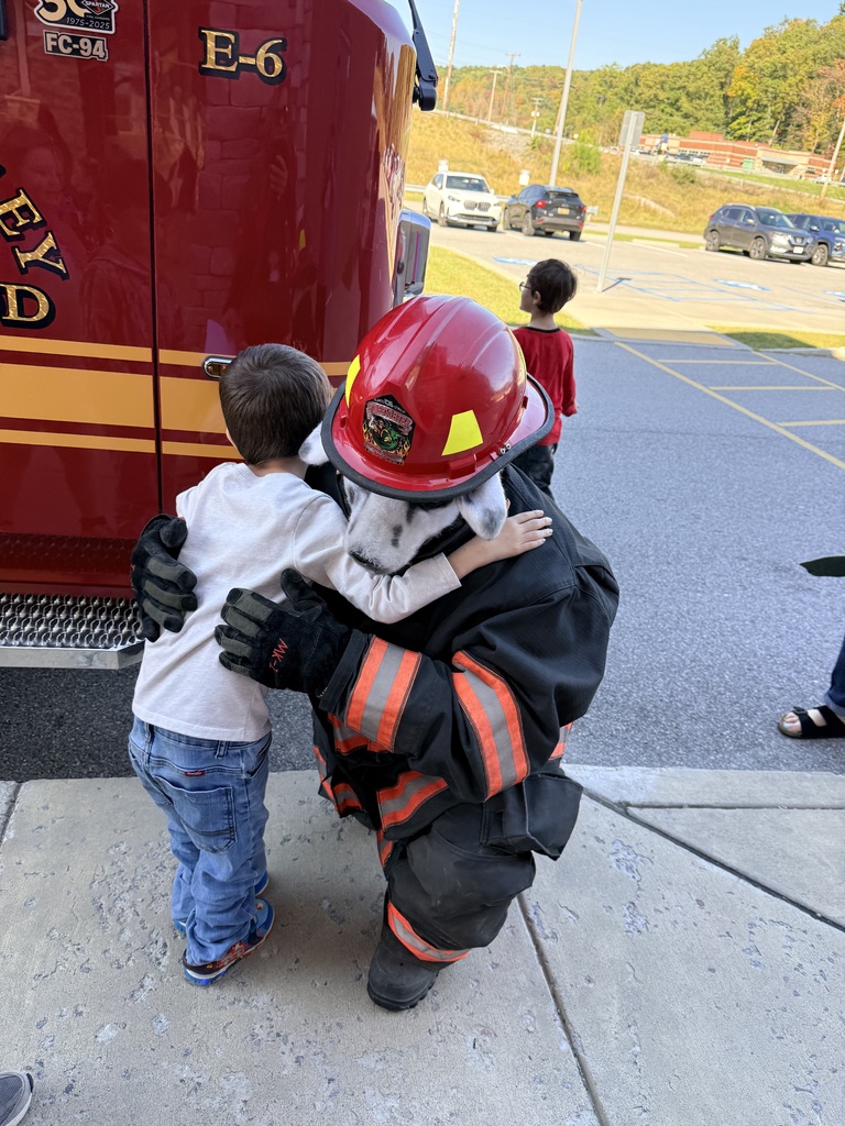 A firefighter in full gear hugs a child in a white t-shirt and jeans near a firetruck.