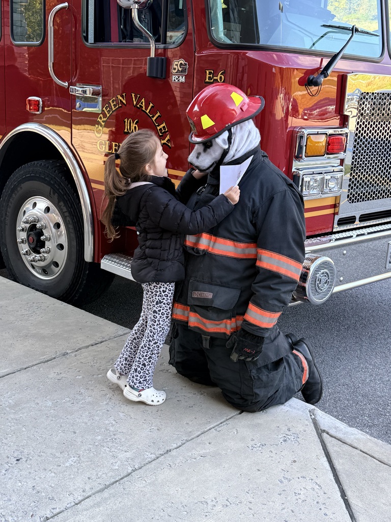 A firefighter kneels in full gear, hugging a young girl in a black jacket. They stand in front of a red firetruck.