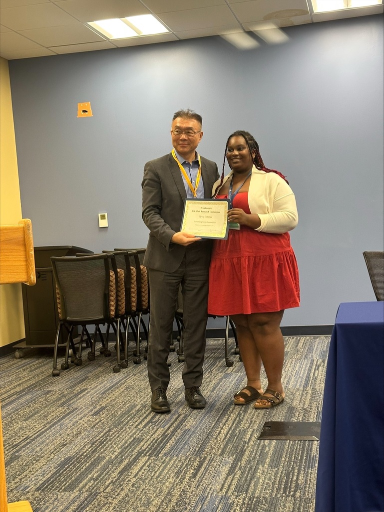A man and a woman in formal attire stand together, holding a framed certificate. Behind them, chairs are arranged, and a table with a blue cloth is visible.