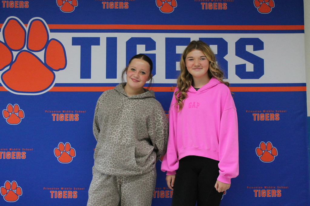 Two girls stand in front of a blue backdrop with the word "Tigers" and paw prints. One wears a gray hoodie; the other wears a pink hoodie.