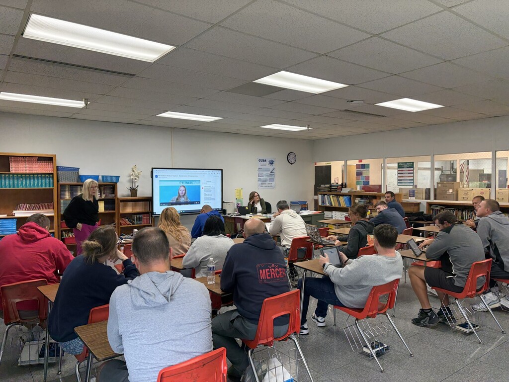 A group of people seated around desks in a classroom. A teacher stands before them near a screen.