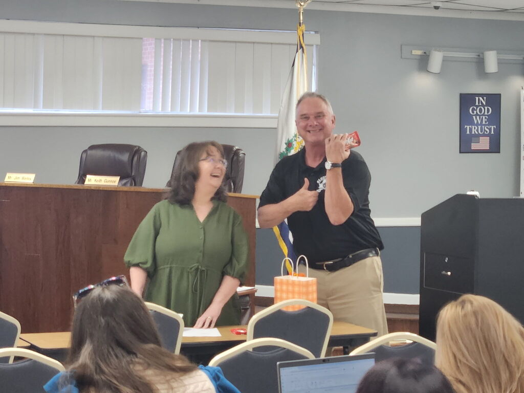Two people in a room; one holds a toy while the other stands beside a table. Behind them is a podium.