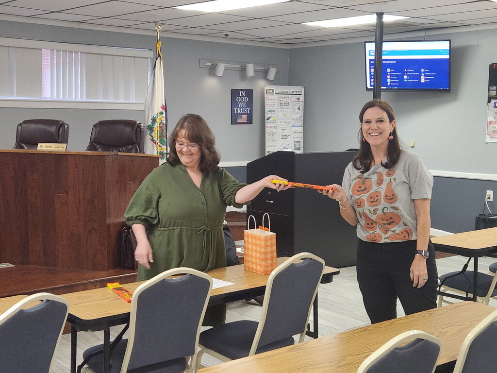 Two women in a conference room with desks and chairs. One holds a carrot and the other a small orange bag.