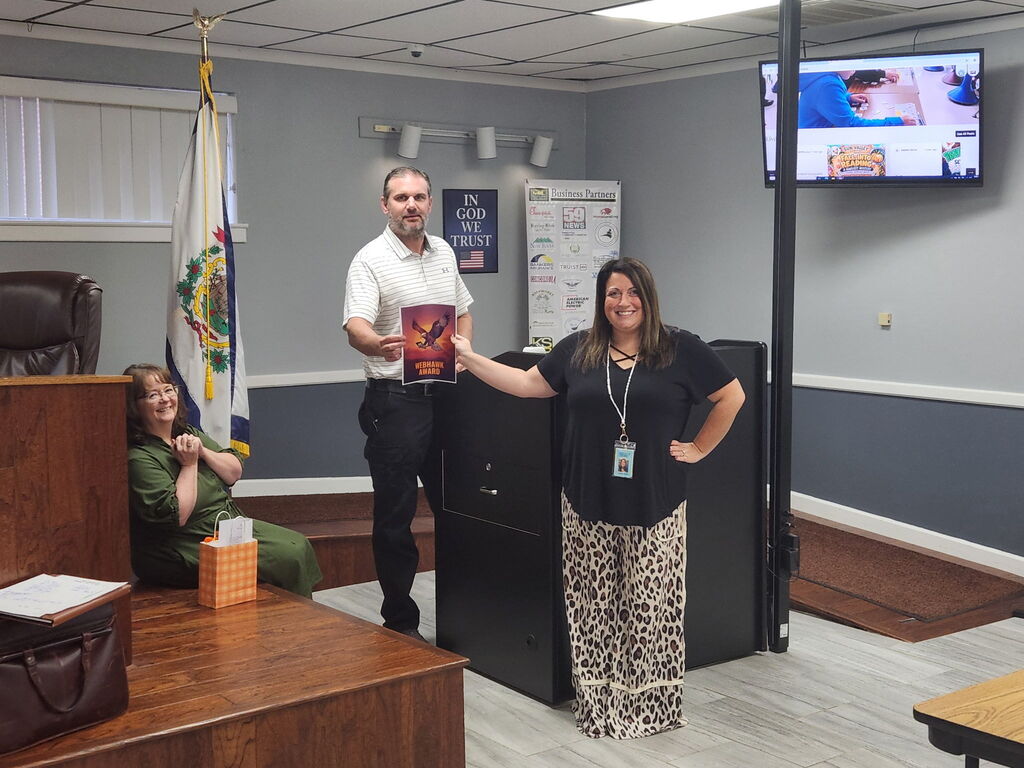 Three people in a room with a TV. One holds a book, another sits in a chair, and a third stands.